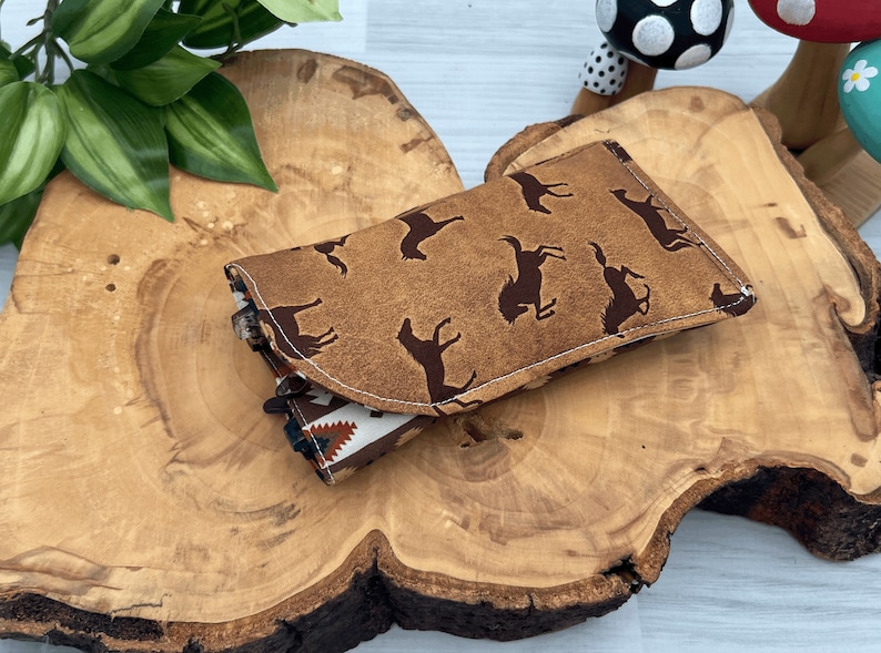 Op de afbeelding: Een bruine brillenkoker met een herhaald patroon van donkerbruine bucking broncos. De koker is open en onthult een patroon aan de binnenkant. Het rust op een houten oppervlak met een natuurlijke rand, met groene bladeren op de achtergrond.