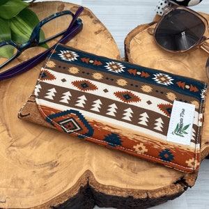 Op de afbeelding: Een stoffen brillenkoker met een zuidwestelijk patroon in bruin, cr&egrave;me en teal. De koker is rechthoekig met een gebogen rand en een label met de tekst "Jeanette Marie". Een bril en zonnebril zijn ook zichtbaar.