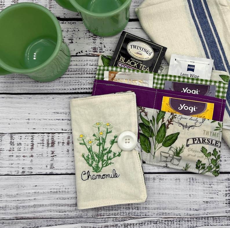 May include: A white fabric pouch with a button closure and embroidered chamomile flower design. The pouch has multiple pockets and is filled with tea bags and other items.