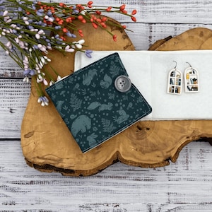 May include: A teal blue fabric wallet with a woodland animal print and a gray button closure. The wallet is sitting on a wooden surface with a white background.