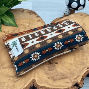 Op de afbeelding: Een rechthoekige, opgevouwen stoffen etui met een patroon ge&iuml;nspireerd op het zuidwesten. Het ontwerp bevat herhaalde geometrische vormen in bruin, wit, blauwgroen en roest. Een wit label met groene bladeren en tekst is op de voorkant genaaid.