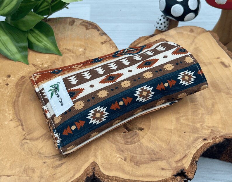 Op de afbeelding: Een rechthoekige, opgevouwen stoffen etui met een patroon ge&iuml;nspireerd op het zuidwesten. Het ontwerp bevat herhaalde geometrische vormen in bruin, wit, blauwgroen en roest. Een wit label met groene bladeren en tekst is op de voorkant genaaid.