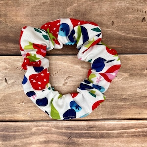 May include: A white scrunchie with a colorful fruit pattern. The pattern includes red strawberries, blue blueberries, green leaves, and pink flowers. The scrunchie is made of fabric and is designed to hold hair.