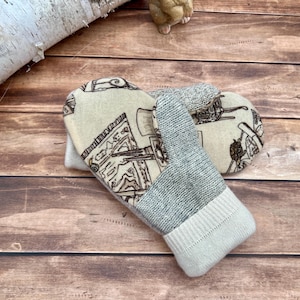 May include: A pair of mittens featuring a cream-colored fabric back with a brown line-art illustration of furniture and household items, paired with a gray knit palm and a solid cream-colored ribbed knit cuff at the wrist opening.