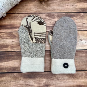May include: A pair of mittens featuring a cream-colored fabric with a brown line-art print on one side and solid heather gray wool on the other. Each mitten has a ribbed cream-colored cuff, with one cuff featuring a single black button.