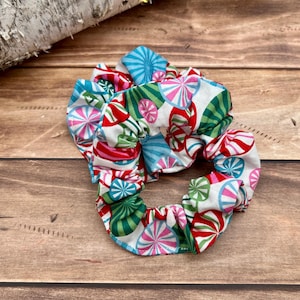 May include: A pair of colorful scrunchies with a candy cane pattern. The scrunchies feature a white background with red, green, pink, and blue candy cane designs. The hair accessories are made of fabric and are suitable for various hairstyles.