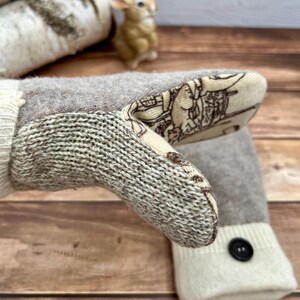 May include: A pair of beige and cream knit mittens featuring a thumb section with a cream-colored fabric overlay printed with a brown line-art illustration. The wrist cuffs are cream-colored knit with a single black button detail on each.