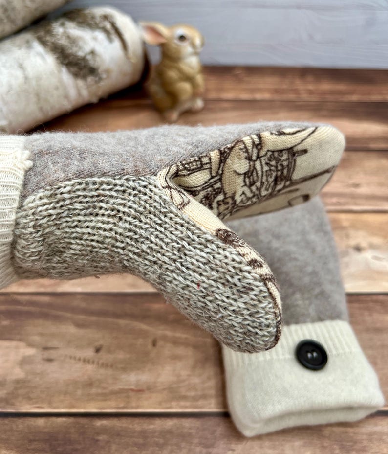 May include: A pair of beige and cream knit mittens featuring a thumb section with a cream-colored fabric overlay printed with a brown line-art illustration. The wrist cuffs are cream-colored knit with a single black button detail on each.