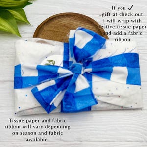 Op de afbeelding: Een cadeau verpakt met een blauw-witte geruite strik. Het cadeau is verpakt in wit vloeipapier met blauw-witte accenten. De tekst geeft aan dat het cadeau wordt verpakt met feestelijk vloeipapier en een stoffen lint.
