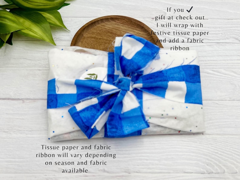 Op de afbeelding: Een cadeau verpakt met een blauw-witte geruite strik. Het cadeau is verpakt in wit vloeipapier met blauw-witte accenten. De tekst geeft aan dat het cadeau wordt verpakt met feestelijk vloeipapier en een stoffen lint.