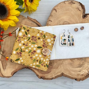 May include: A handmade jewelry display featuring a mustard yellow fabric-covered book-style case with a mushroom and floral pattern. A white fabric jewelry holder displays two pairs of earrings: window-shaped dangle earrings and small stud earrings. Sunflowers and berries are in the background.