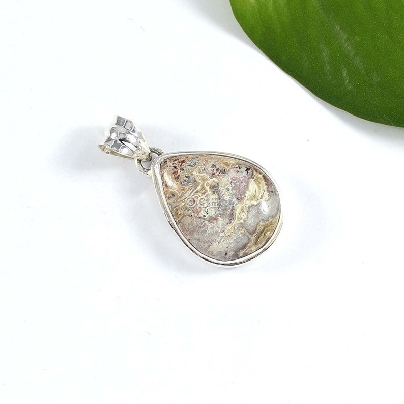 Puede incluir: Un colgante de plata con una piedra de &aacute;gata de color marr&oacute;n y blanco en forma de l&aacute;grima. La piedra tiene un patr&oacute;n de remolino y est&aacute; engastada en un bisel de plata.