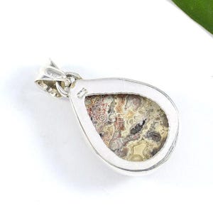 Puede incluir: Un colgante de plata con una piedra en forma de l&aacute;grima que presenta un patr&oacute;n de &aacute;gata colorido. La piedra est&aacute; engastada en un bisel de plata y tiene un aro de suspensi&oacute;n de plata.