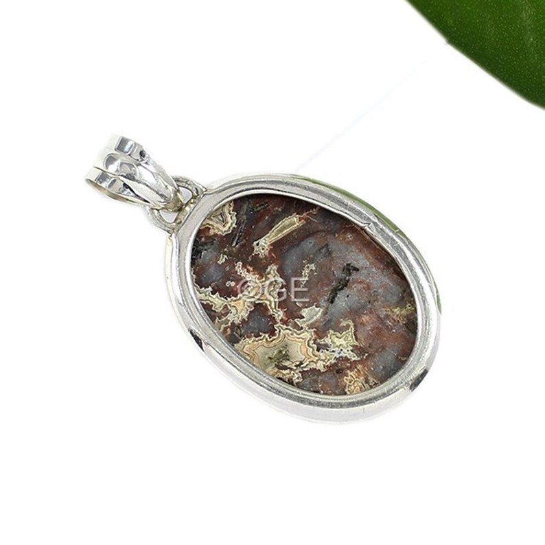 Puede incluir: Pendiente de plata con una piedra de &aacute;gata marr&oacute;n, blanca y gris con un patr&oacute;n de remolino. La piedra est&aacute; engastada en un marco ovalado de plata.