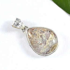Puede incluir: Un colgante de plata con una piedra de &aacute;gata en forma de l&aacute;grima de color marr&oacute;n, blanco y rosa.