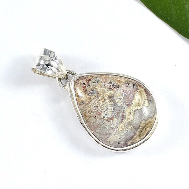 Puede incluir: Un colgante de plata con una piedra de &aacute;gata en forma de l&aacute;grima de color marr&oacute;n, blanco y rosa.