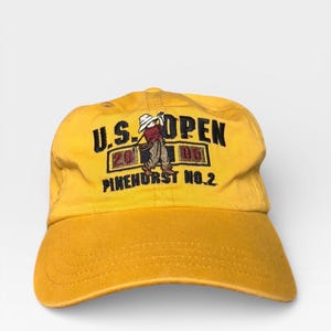 May include: Yellow baseball cap with embroidered text and a golf-themed graphic. The text reads "U.S. OPEN 2005 PINEHURST NO. 2" in black. The graphic depicts a golfer in action. The cap has a curved brim and adjustable strap.