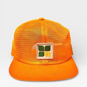 Può includere: Cappellino da camionista arancione brillante con retro in rete e visiera curva. Il cappello presenta una toppa quadrata con un design verde e giallo. Il cappello è di colore arancione vibrante.