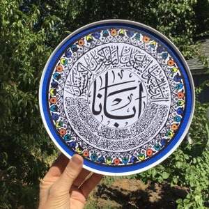 May include: A round, white plate with a blue border and colorful floral designs. The center of the plate features a black and white Arabic calligraphy design with the word "Allah" in the center.
