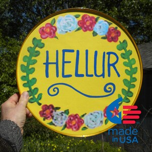 May include: A yellow round sign with a gold trim and a floral border. The sign says "HELLUR" in blue letters with a blue swirl design. The sign also has a red, white, and blue star and the words "made in USA" in black letters.