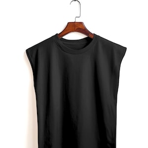 Puede incluir: Una camiseta negra sin mangas lisa colgada de una percha de madera. La camiseta tiene cuello redondo y un diseño sencillo y minimalista. El fondo es blanco, lo que resalta el color y la forma de la camiseta.