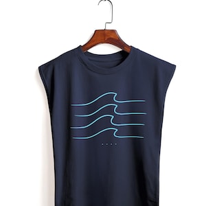 Könnte beinhalten: Marineblaues ärmelloses T-Shirt mit hellblauem Wellenmuster. Die Grafik zeigt vier geschwungene Linien, die Wellen darstellen, mit drei kleinen Punkten darunter. Das T-Shirt hängt an einem Holzbügel.