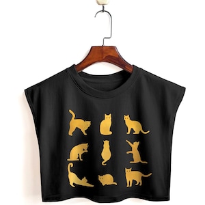 Op de afbeelding: Zwarte mouwloze crop top met een gouden kattenpatroon. De katten staan in verschillende poses, wat een speels ontwerp creëert. De top hangt aan een houten hanger tegen een witte achtergrond.