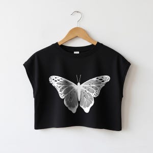 Könnte beinhalten: Ein schwarzes bauchfreies T-Shirt mit kurzen Ärmeln und Rundhalsausschnitt, das in der Mitte mit einem großen weißen Schmetterling bedruckt ist, der komplexe Flügelmuster und kleine schwarze Sprenkel aufweist, präsentiert auf einem Holzkleiderbügel.