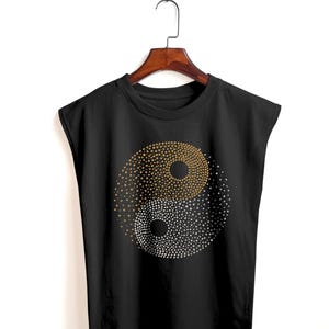 May include: Black sleeveless top with a gold and white dotted Yin Yang design. The Yin Yang symbol is the focal point, with the two halves distinguished by colour and dot patterns. The top is hanging on a wooden hanger.