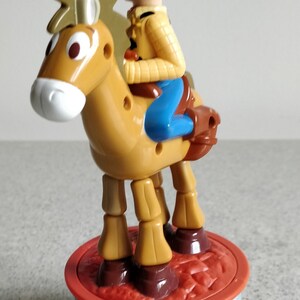 K&ouml;nnte beinhalten: Ein Plastikspielzeug von Woody aus Toy Story, das auf einem braunen Pferd reitet. Woody tr&auml;gt seinen charakteristischen braunen Hut und eine blaue Jeans. Das Pferd hat einen braunen Sattel und steht auf einem roten Sockel mit einem Cartoon-Bild einer Westernstadt.