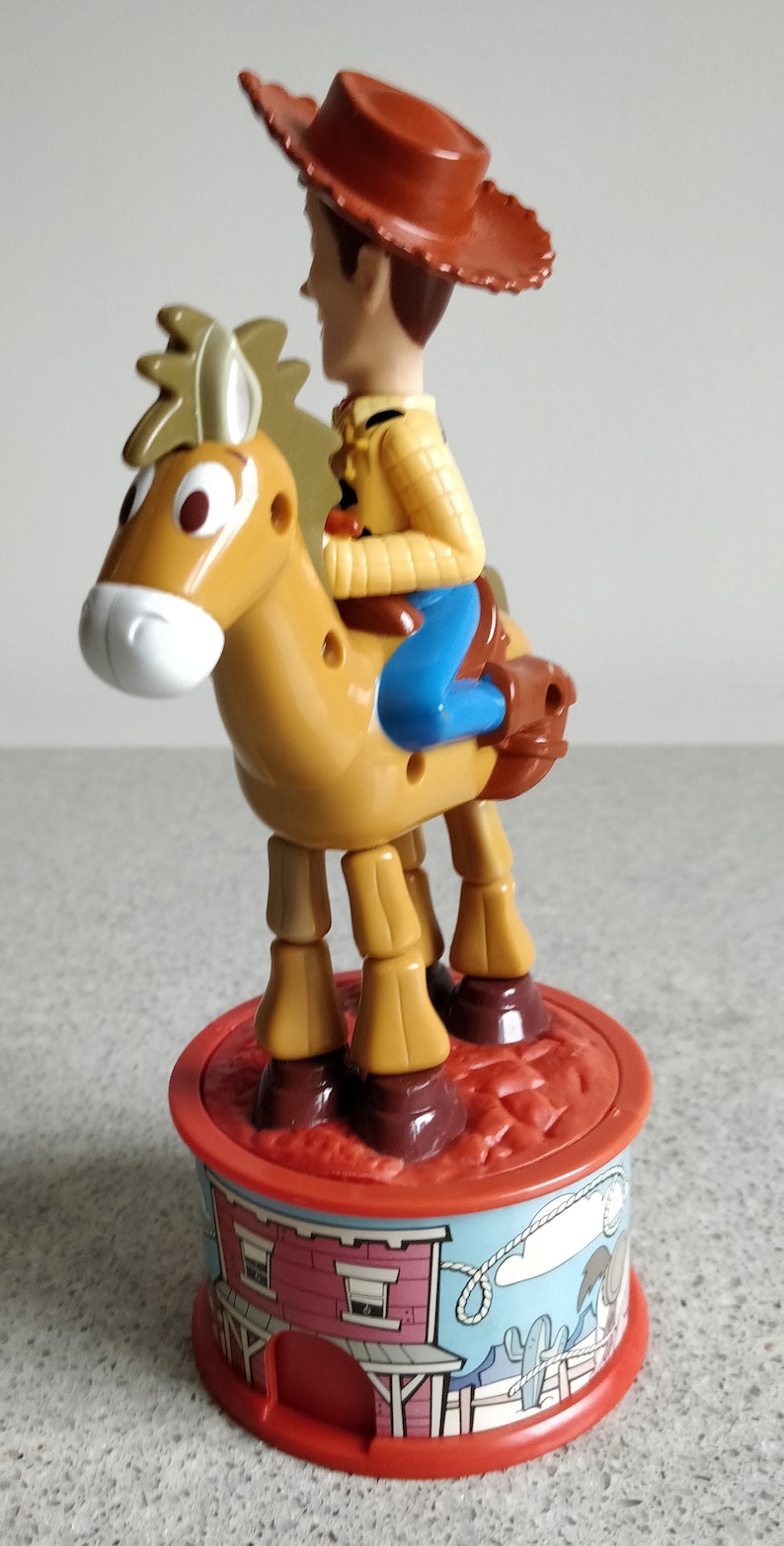 K&ouml;nnte beinhalten: Ein Plastikspielzeug von Woody aus Toy Story, das auf einem braunen Pferd reitet. Woody tr&auml;gt seinen charakteristischen braunen Hut und eine blaue Jeans. Das Pferd hat einen braunen Sattel und steht auf einem roten Sockel mit einem Cartoon-Bild einer Westernstadt.