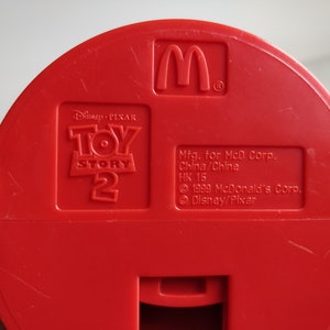 K&ouml;nnte beinhalten: Roter Plastikspielzeugbeh&auml;lter mit dem Toy Story 2 Logo und dem goldenen M von McDonald's. Der Beh&auml;lter tr&auml;gt den Text "Mfg. for McD Corp. China/China HK 15 &copy; 1999 McDonald's Corp. &copy; Disney/Pixar".