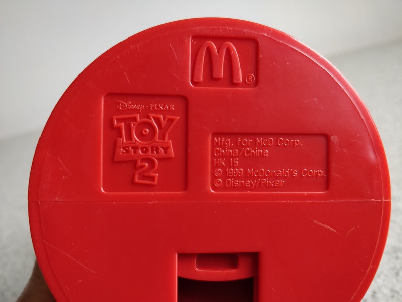 K&ouml;nnte beinhalten: Roter Plastikspielzeugbeh&auml;lter mit dem Toy Story 2 Logo und dem goldenen M von McDonald's. Der Beh&auml;lter tr&auml;gt den Text "Mfg. for McD Corp. China/China HK 15 &copy; 1999 McDonald's Corp. &copy; Disney/Pixar".