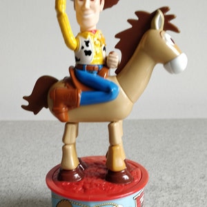 K&ouml;nnte beinhalten: Ein Plastikspielzeug von Woody aus Toy Story, der auf seinem Pferd Bullseye reitet. Woody tr&auml;gt seinen charakteristischen Cowboyhut, seine Weste und seine Jeans. Die Basis des Spielzeugs ist rot und zeigt ein Cartoon-Bild von Woody und Bullseye.