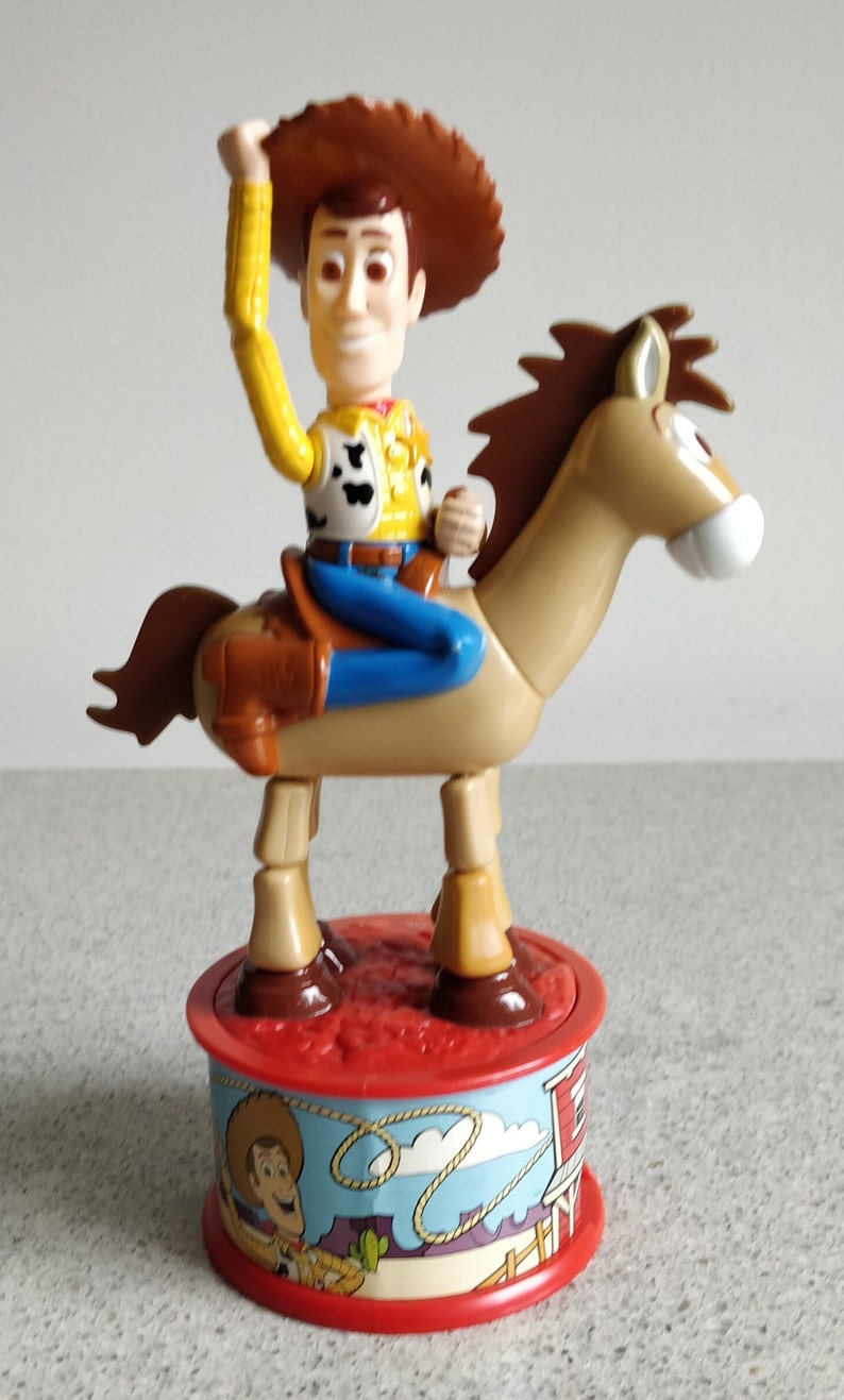 K&ouml;nnte beinhalten: Ein Plastikspielzeug von Woody aus Toy Story, der auf seinem Pferd Bullseye reitet. Woody tr&auml;gt seinen charakteristischen Cowboyhut, seine Weste und seine Jeans. Die Basis des Spielzeugs ist rot und zeigt ein Cartoon-Bild von Woody und Bullseye.
