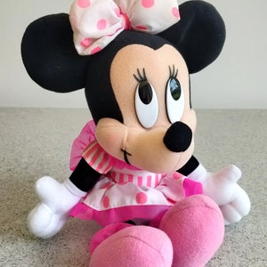 Puede incluir: Un juguete de peluche de Minnie Mouse en blanco y negro con un lazo rosa y blanco con lunares. El juguete lleva un vestido a rayas rosa y blanco con lunares rosas. El juguete est&aacute; sentado con las piernas cruzadas y los brazos extendidos.