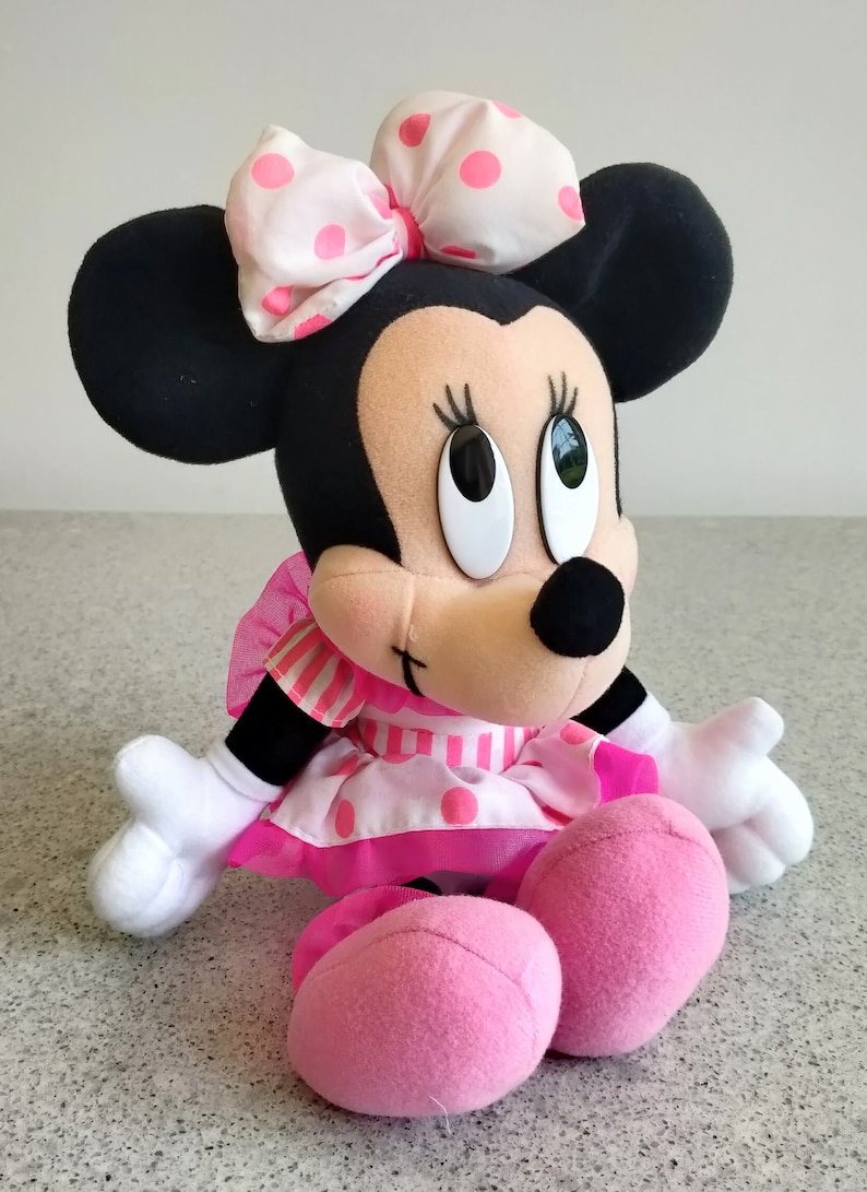 Puede incluir: Un juguete de peluche de Minnie Mouse en blanco y negro con un lazo rosa y blanco con lunares. El juguete lleva un vestido a rayas rosa y blanco con lunares rosas. El juguete est&aacute; sentado con las piernas cruzadas y los brazos extendidos.