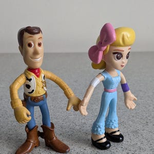May include: Toy Story characters Woody and a girl with a pink bow in her hair. Woody is wearing his signature cowboy hat, vest, and jeans. The girl is wearing a blue dress with a pink belt and purple wristbands.