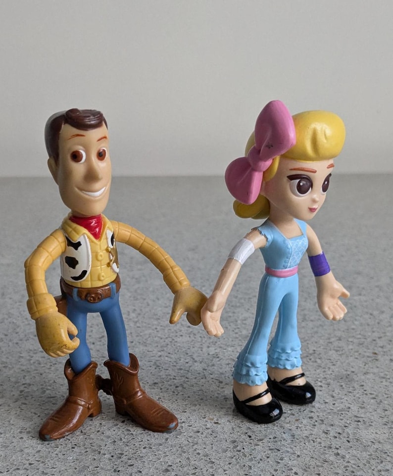 May include: Toy Story characters Woody and a girl with a pink bow in her hair. Woody is wearing his signature cowboy hat, vest, and jeans. The girl is wearing a blue dress with a pink belt and purple wristbands.