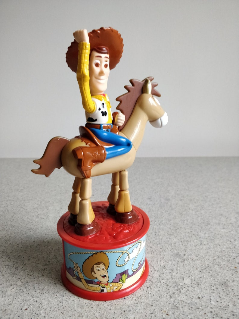 K&ouml;nnte beinhalten: Ein Plastikspielzeug von Woody aus Toy Story, das auf einem braunen Pferd reitet. Woody tr&auml;gt seinen charakteristischen Cowboyhut, seine Weste und seine Jeans. Das Spielzeug steht auf einem roten Sockel mit einem Bild von Woody darauf.