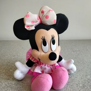Puede incluir: Un juguete de peluche de Minnie Mouse con un vestido de lunares rosa y blanco y un lazo rosa y blanco con lunares. El juguete tiene orejas negras y una nariz rosa.