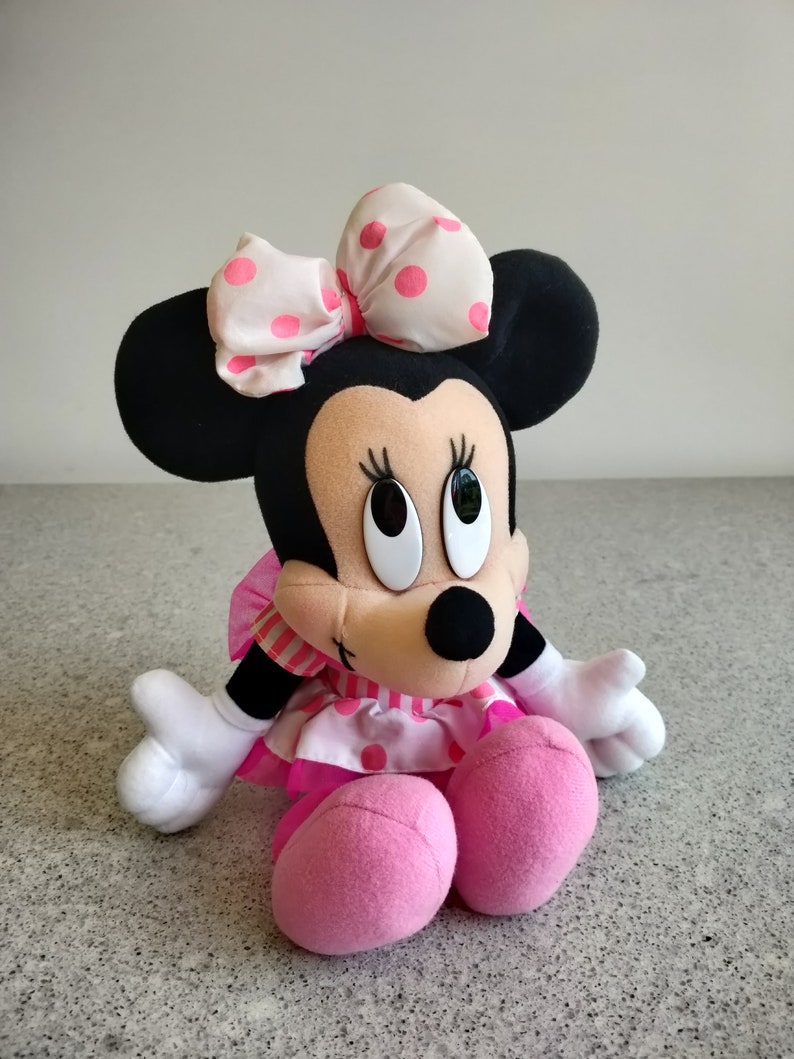 Puede incluir: Un juguete de peluche de Minnie Mouse con un vestido de lunares rosa y blanco y un lazo rosa y blanco con lunares. El juguete tiene orejas negras y una nariz rosa.