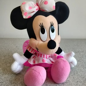Puede incluir: Un juguete de peluche de Minnie Mouse con un vestido de lunares rosa y blanco y un lazo blanco con lunares rosas. Tiene grandes ojos negros y una nariz negra.