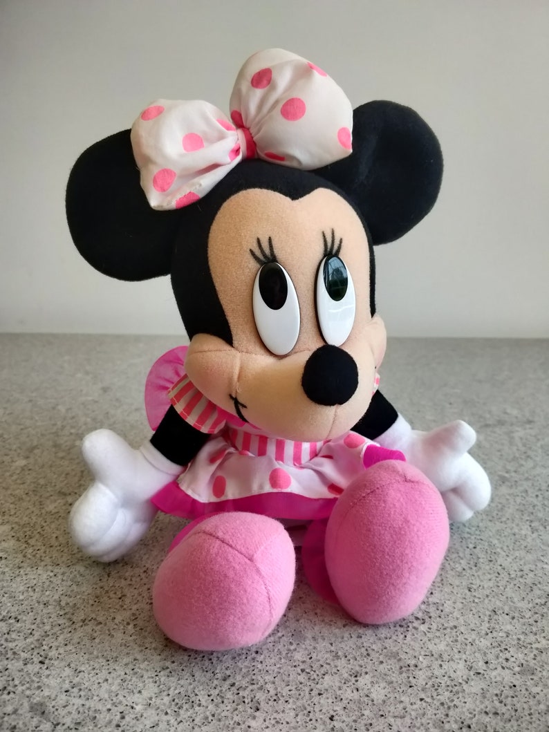 Puede incluir: Un juguete de peluche de Minnie Mouse con un vestido de lunares rosa y blanco y un lazo blanco con lunares rosas. Tiene grandes ojos negros y una nariz negra.