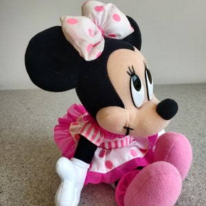 Puede incluir: Un juguete de peluche de Minnie Mouse con un vestido rosa y blanco de lunares y un lazo rosa. El juguete est&aacute; sentado en una superficie gris.