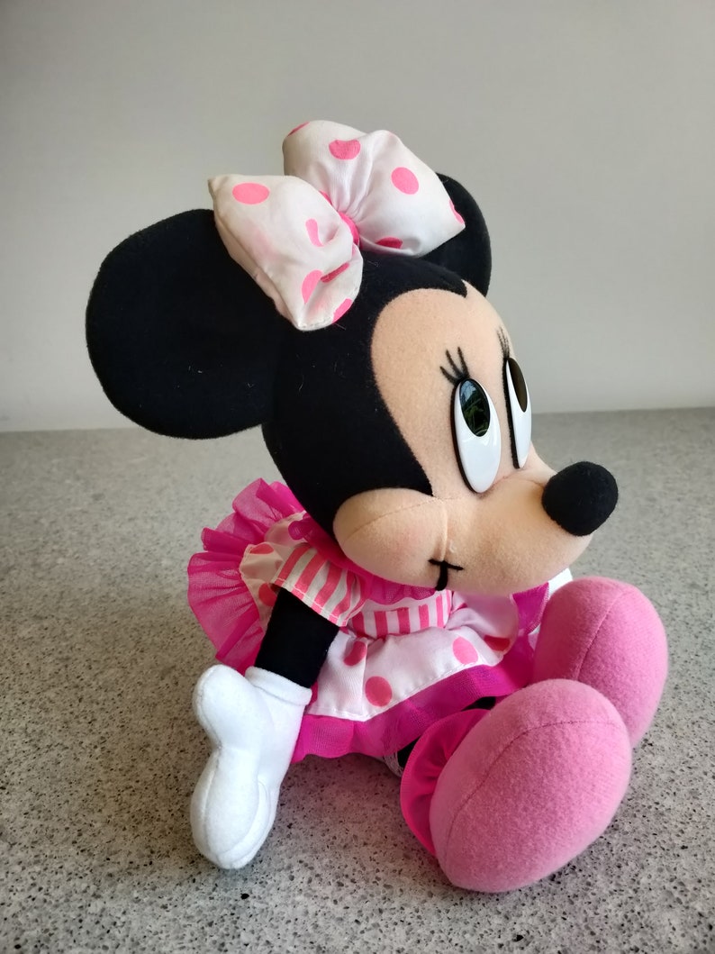 Puede incluir: Un juguete de peluche de Minnie Mouse con un vestido rosa y blanco de lunares y un lazo rosa. El juguete est&aacute; sentado en una superficie gris.