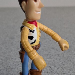 May include: A plastic toy figure of Woody from the Toy Story franchise. Woody is wearing his signature brown cowboy hat, yellow shirt, blue jeans, and brown cowboy boots. He has a friendly smile and brown hair.