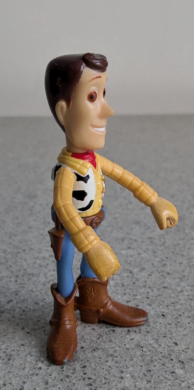 May include: A plastic toy figure of Woody from the Toy Story franchise. Woody is wearing his signature brown cowboy hat, yellow shirt, blue jeans, and brown cowboy boots. He has a friendly smile and brown hair.