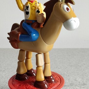 K&ouml;nnte beinhalten: Ein Plastikspielzeug von Woody aus Toy Story, das auf einem braunen Pferd reitet. Woody tr&auml;gt seinen charakteristischen Cowboyhut, seine Weste und seine Jeans. Das Pferd hat bewegliche Beine und steht auf einem roten und blauen Sockel mit einer Western-Szene.