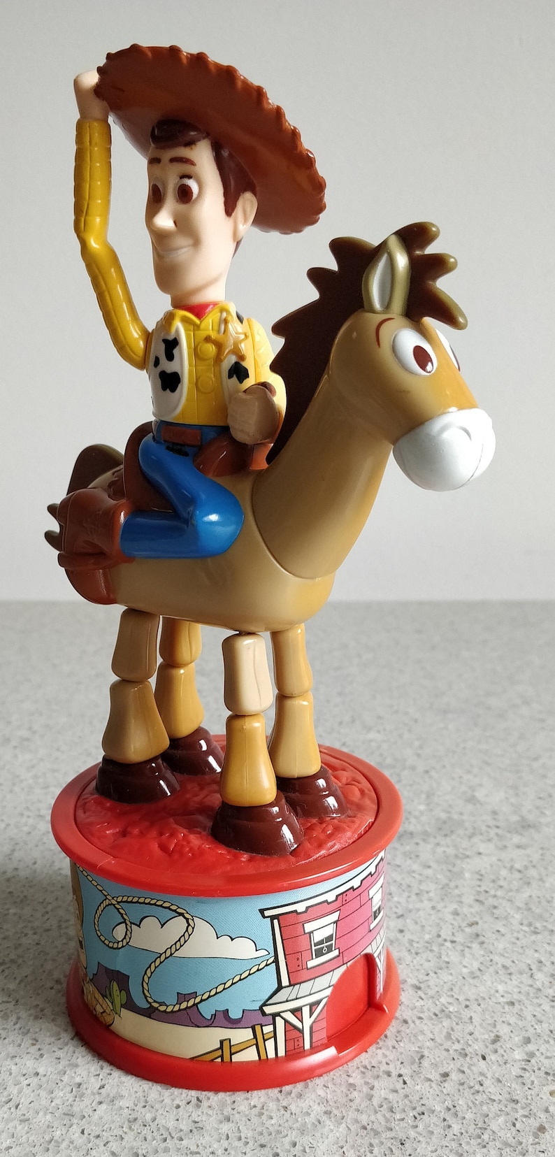 K&ouml;nnte beinhalten: Ein Plastikspielzeug von Woody aus Toy Story, das auf einem braunen Pferd reitet. Woody tr&auml;gt seinen charakteristischen Cowboyhut, seine Weste und seine Jeans. Das Pferd hat bewegliche Beine und steht auf einem roten und blauen Sockel mit einer Western-Szene.